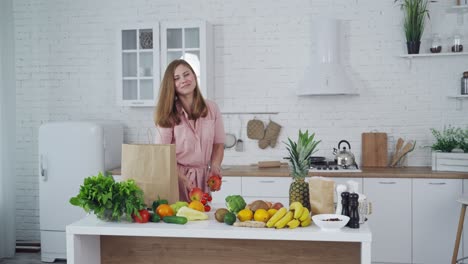 Happy young woman with fresh apples in the kitchen. Beautiful housewife holds and sniffs natural fruit on the background of healthy food on the table.
