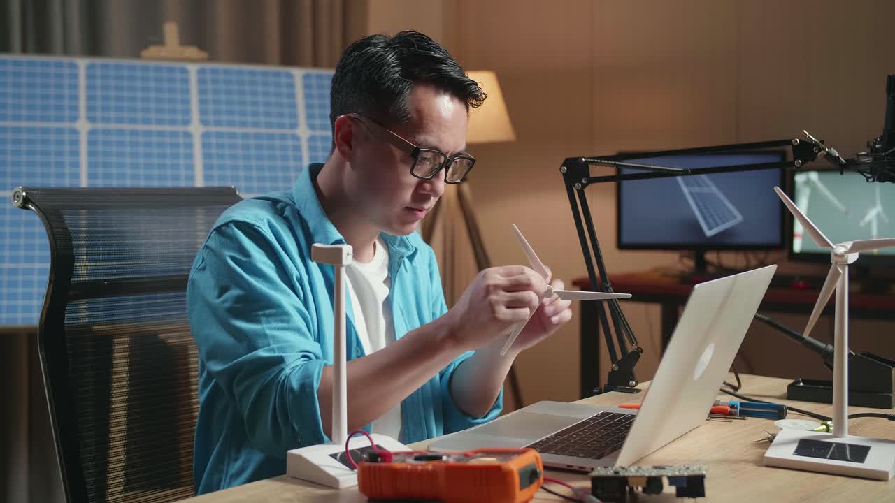 hombre asiático ensambla la turbina eólica mientras trabaja con una computadora portátil junto a la célula solar en la oficina