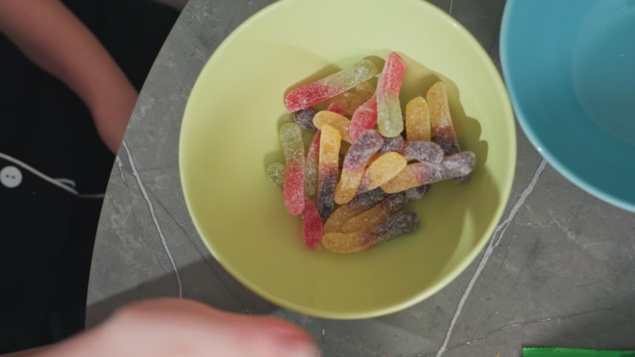 aerial view of people pouring colorful gummy candy from green packs into yellow bowl on kitchen table, blue bowls and green plant visible, casual home setting, lively snack preparation