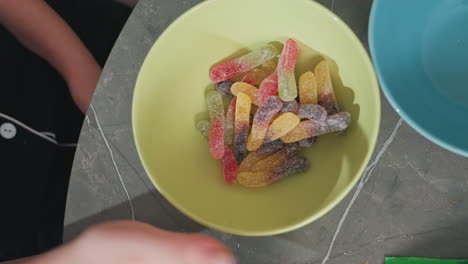 aerial view of people pouring colorful gummy candy from green packs into yellow bowl on kitchen table, blue bowls and green plant visible, casual home setting, lively snack preparation
