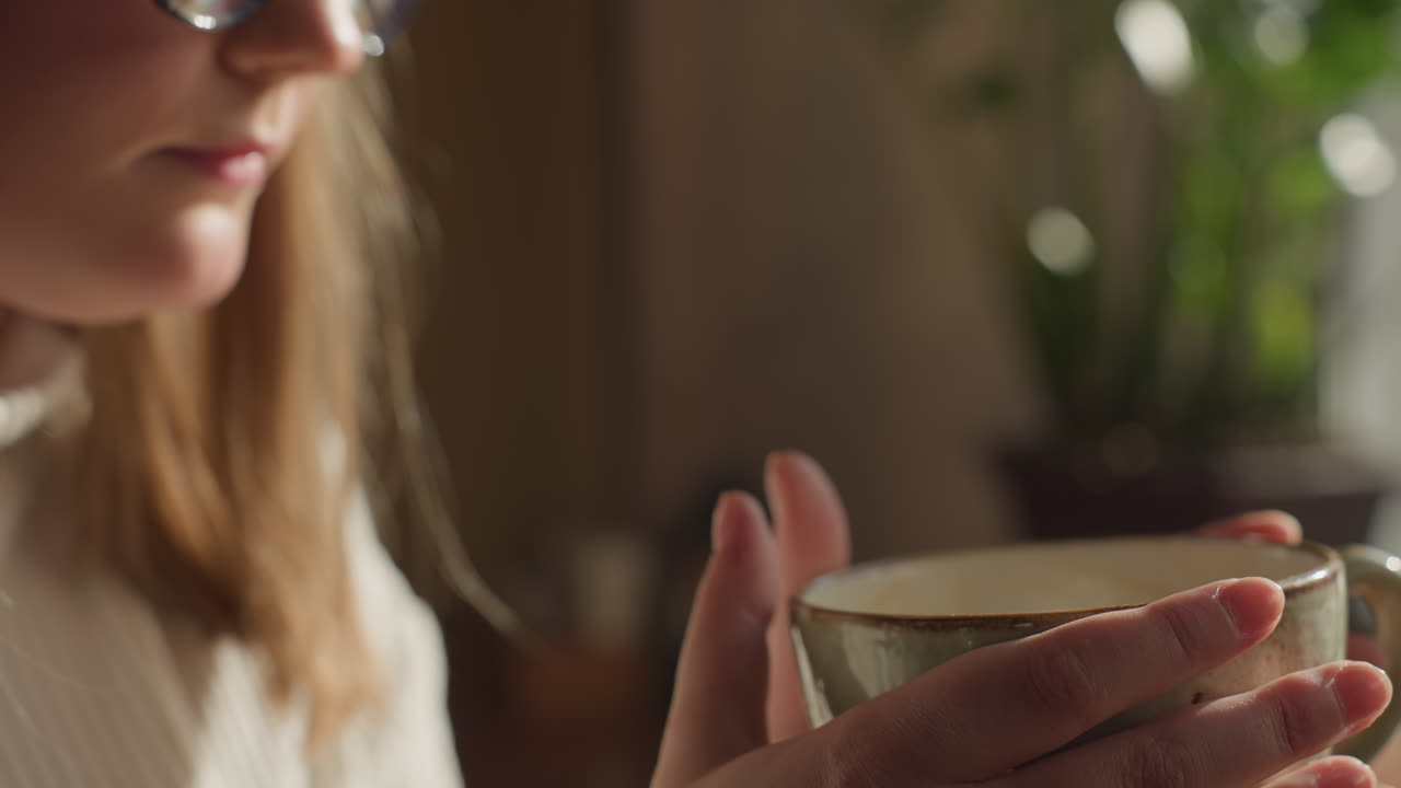 Close up of fair skin woman in ribbed white sweater lifting tea cup slowly while gazing through window with thoughtful expression, soft sunlight illuminating hand and cup, warm tones fill the scene