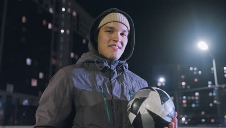 young man in hoodie smiling while skillfully spinning football in his hand during nighttime on an urban sports field surrounded by illuminated buildings and streetlights