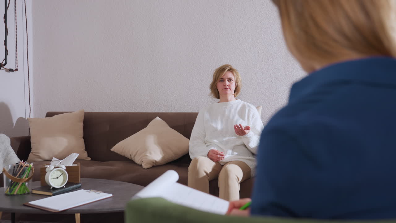 Reflective woman gesturing with her hands while speaking to therapist taking notes in cozy counseling room with brown sofa, pillow, green accent wall, table with clock and pen pack visible