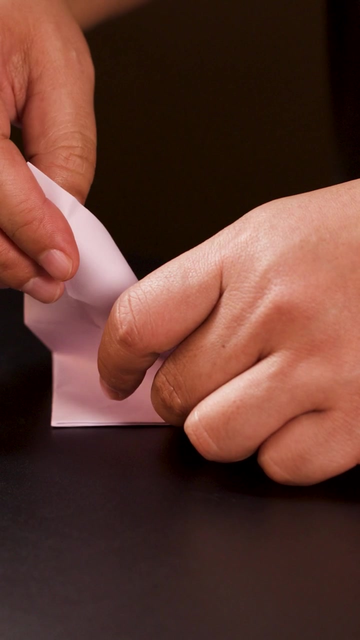 Close-up vertical shot of hands precisely folding white paper into an origami boat shape.