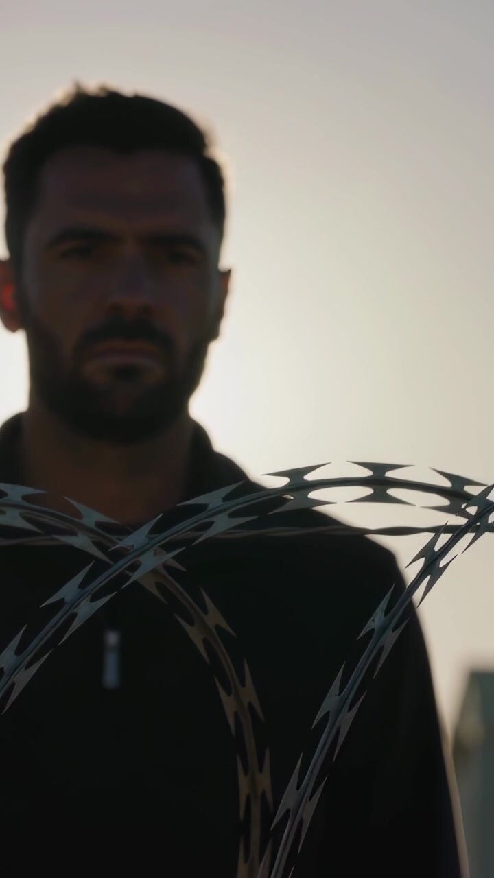 Video verticale: Uomo in giacca di fronte all'obiettivo, in piedi su un tetto, il sole proietta un'ombra dietro il filo spinato.