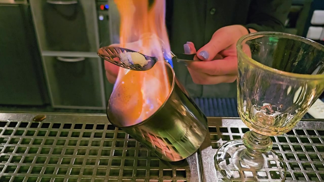 Primo piano suggestivo della preparazione di un cocktail infuocato dietro un bancone, con fiamme blu, mani del barista, una zolletta di zucchero, un cucchiaio forato, una tazza di metallo, un calice di vetro intagliato e una luce soffusa da lounge.