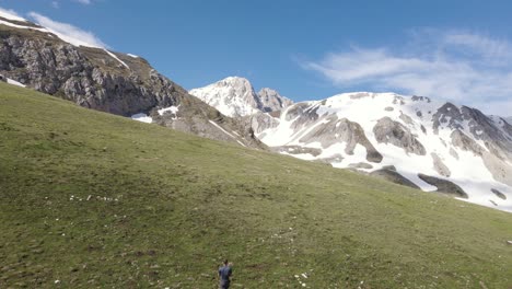 Drone-shot-going-upwards-of-a-young-man-hiking-alone-walking-towards-a-massive-mountain-peak-in-the-rural-countryside-of-Abruzzo-in-Italy