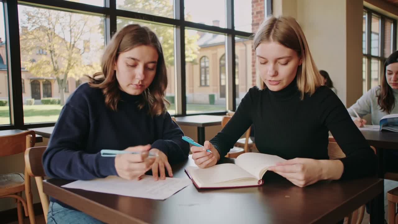 Starting two students studying, writing and consulting red notebook in commons with blue pens