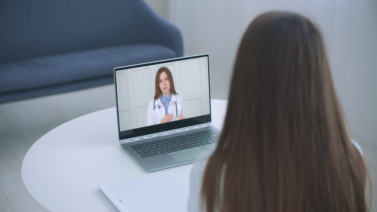 Giovane donna, vista di spalle, in consultazione online tramite videochiamata su laptop con un medico di famiglia o un terapeuta, dopo aver avvertito i primi sintomi di una malattia virale, in un contesto di assicurazione medica e diffusione del Covid-19