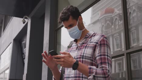 Vista-En-ángulo-Bajo-De-Un-Hombre-Caucásico-Usando-Su-Teléfono-Y-Usando-Una-Máscara-De-Coronavirus-Covid19