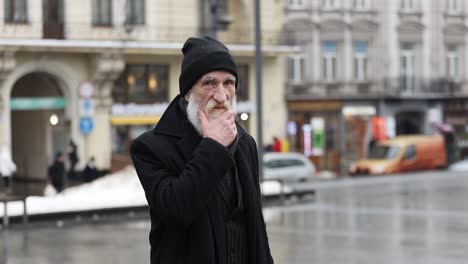 An Elderly Man is Depicted in an Urban Setting Showcasing his Distinctive and Striking White Beard an Elderly Man Adorned with a White Beard and Dressed in all Black Clothing Stands in a Lively Urban Environment