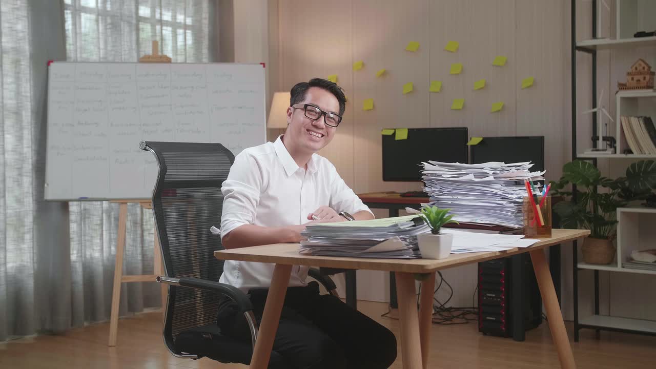 Asian Man Smiling To Camera While Working With Documents At The Office
