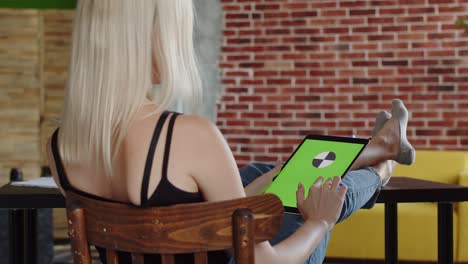 woman is sitting chair with her feet on table and swipe her finger on screen of tablet with chromakey, back view. concept of free time and rest