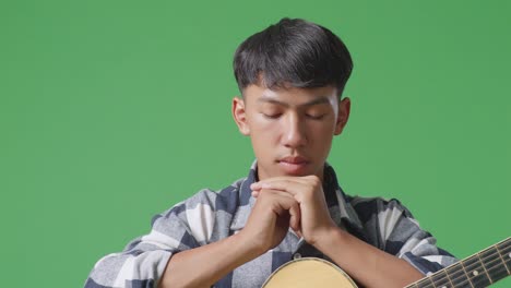 primer plano de un adolescente asiático con una guitarra orando por algo en el fondo de la pantalla verde