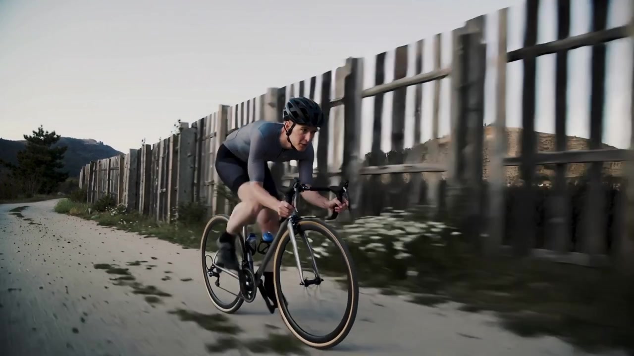 Telecamera che segue un ciclista con una maglia grigia che pedala su una bici da gravel lungo un sentiero sterrato, sollevando sassi