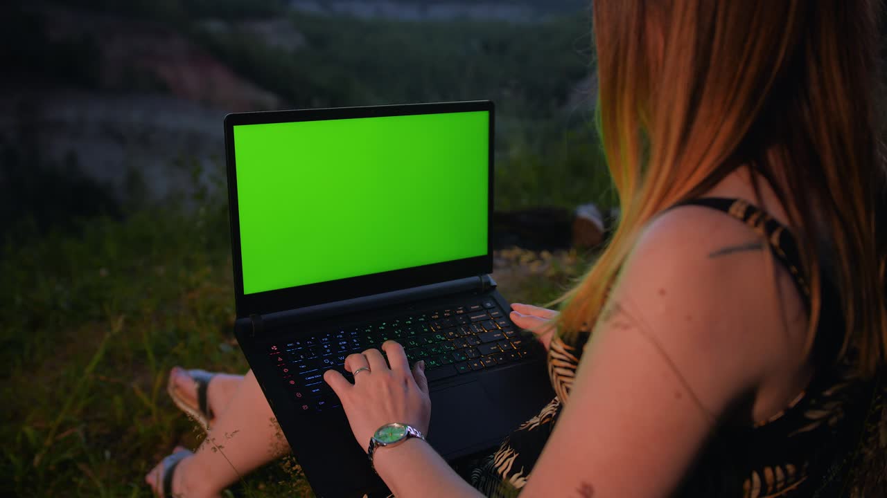 una joven trabaja en una computadora portátil con una pantalla verde en la cima de una montaña.