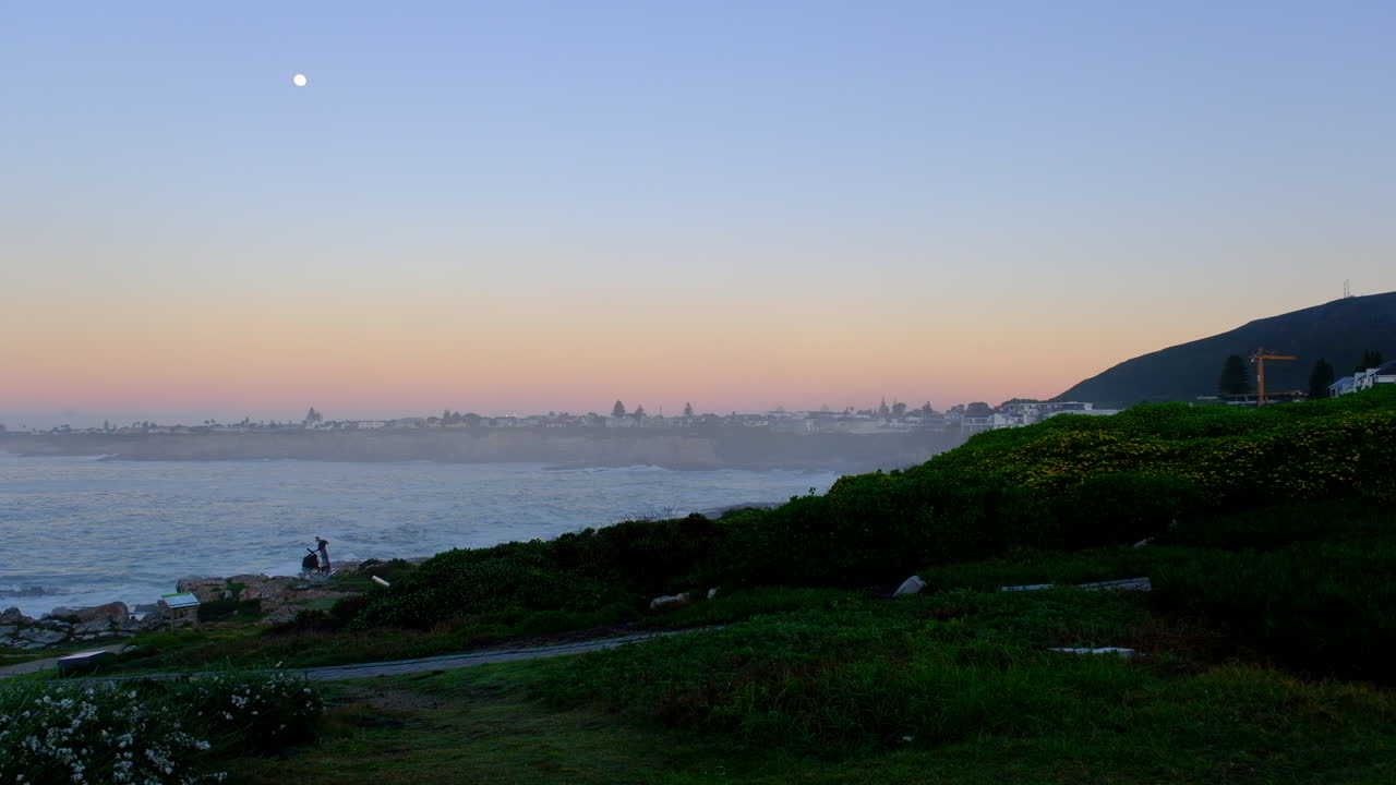 Orange horizon of Hermanus during sunrise with full moon setting, coastline view