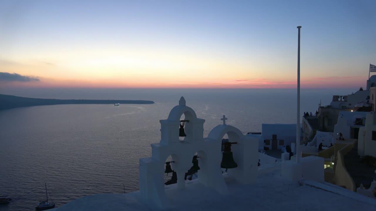 A breathtaking aerial view captures the serene sunset over Santorini, Greece. The iconic white bell towers stand against the vibrant horizon, creating a tranquil, picturesque scene.