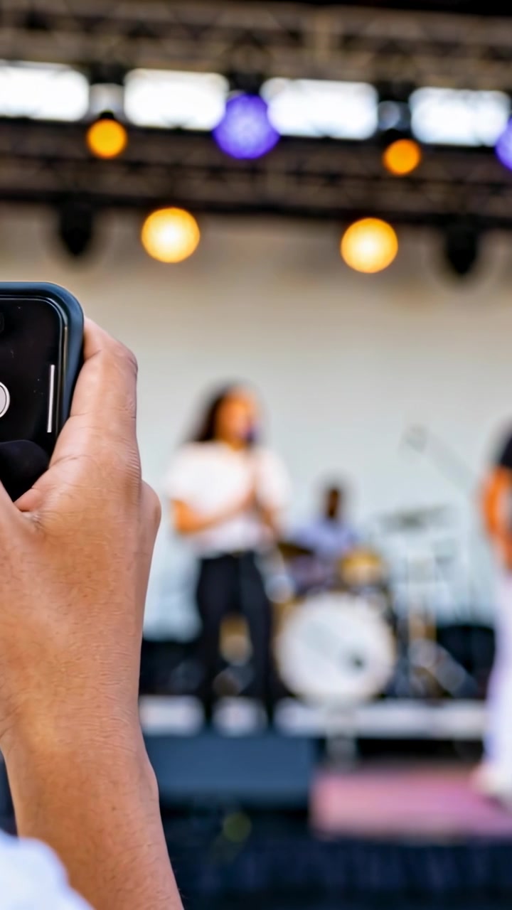 Vertical video: Band playing, hand filming singer in white top on stage phone drums mic, copy space