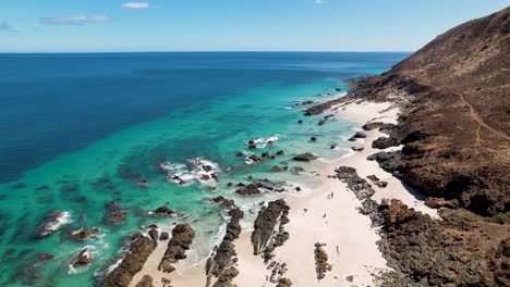 Drohne-Fliegt-Hoch-über-Einem-Wunderschönen-Weißen-Sandstrand-Mit-Felsformationen,-Die-Aus-Dem-Kristallklaren-Blauen-Ozean-Ragen