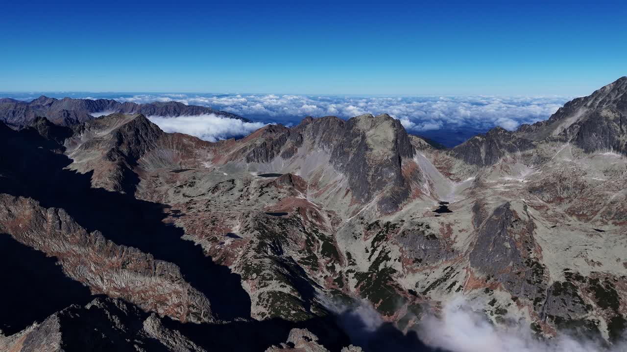 Aerial view of rugged High Tatras mountain peaks rising above a sea of clouds under a clear blue sky. Rocky ridges and autumn tones dominate the scene. Location: High Tatras, Slovakia