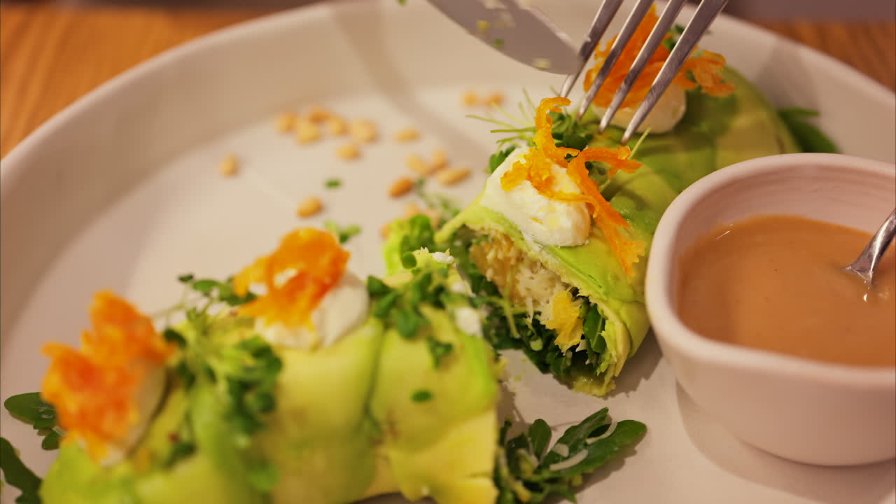 Close up of a woman cutting up an avocado roll with sauce on a white plate at a restaurant
