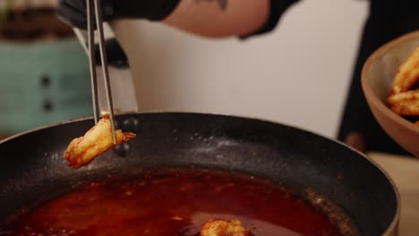 captura de pantalla de un chef profesional usando pinzas de acero inoxidable, sujetando un langostino pelado cocido recubierto con jugosa salsa de paprika roja de una sartén caliente en un bol a cámara lenta