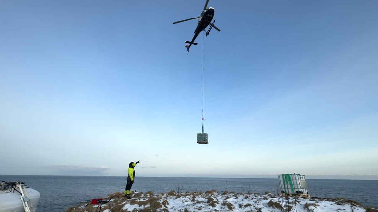Medium shot of a helicopter bringing in supplies on a longline near the ocean with an operator directing and ready to receive