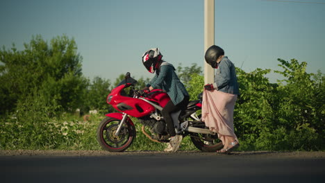 two women with helmets, one sitting on a red sports bike preparing to move, while the other lady lifts her gown as she climb the bike