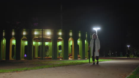 woman dressed in white coat holding umbrella walking through urban park at night, glowing green pavilion illuminated in background under bright streetlights, other people visible in distance along pathway