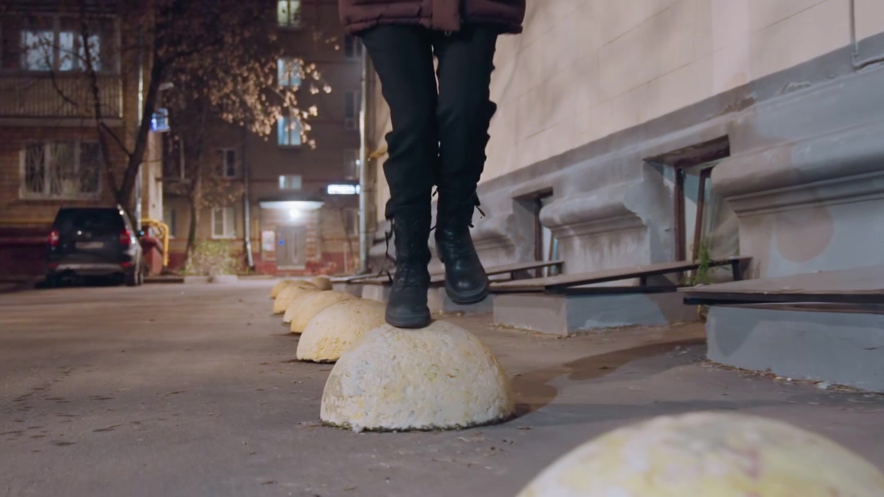 Urban alley walker hopping over bollards, narrow street scene with worn yellow domes, confident black boots stepping in rhythm, casual bag swing, lamplight reflections on asphalt, quick sequence