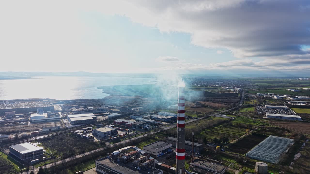 Smoke rises from a tall chimney near a coastal industrial area in daylight