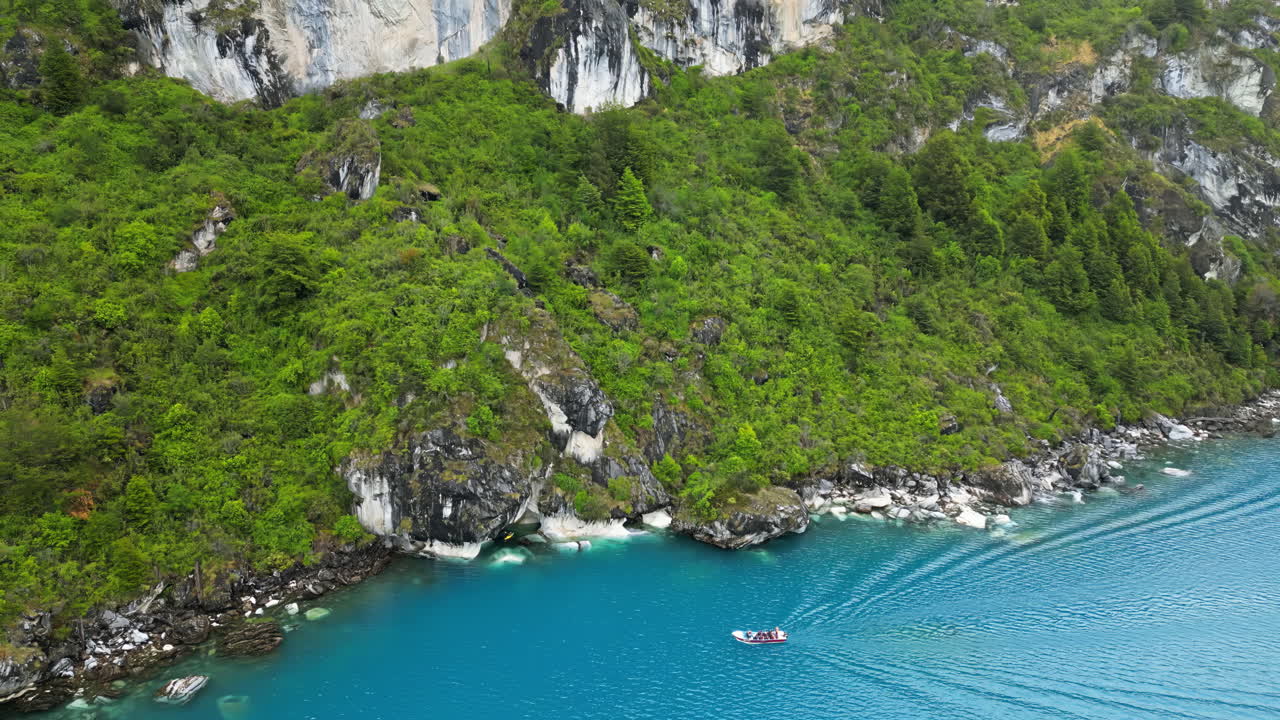Aerial drone view of a small boat gliding across a bright turquoise lake surrounded by forested cliffs in Patagonia