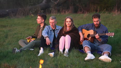group of friends enjoying summer evening in the nature. young men playing music and singing songs. company sitting near the campfire.