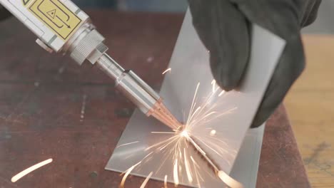 man with gloves showcasing laser welding device, connecting two rectangular pieces of sheet metal while sparks fly