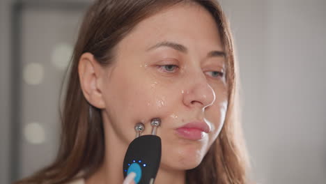 woman does microcurrent procedure for face lifting. pretty brunette woman runs hand with electric device over face in home bathroom slow motion