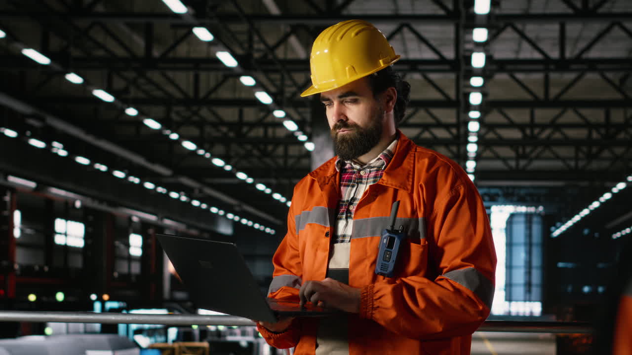 Ingegnere al laptop, che esamina sistemi meccanici, in un enorme capannone industriale.