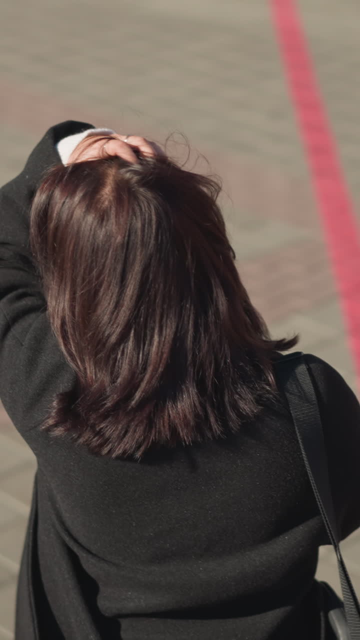 밖에서 고 머리를 만지고 손에 드폰을 들고 있는 여성이 활기찬 환경으로 도시 보행로를 따라 걸어가는 모습이 뒷면에서 명확하게 포착되어 있습니다.