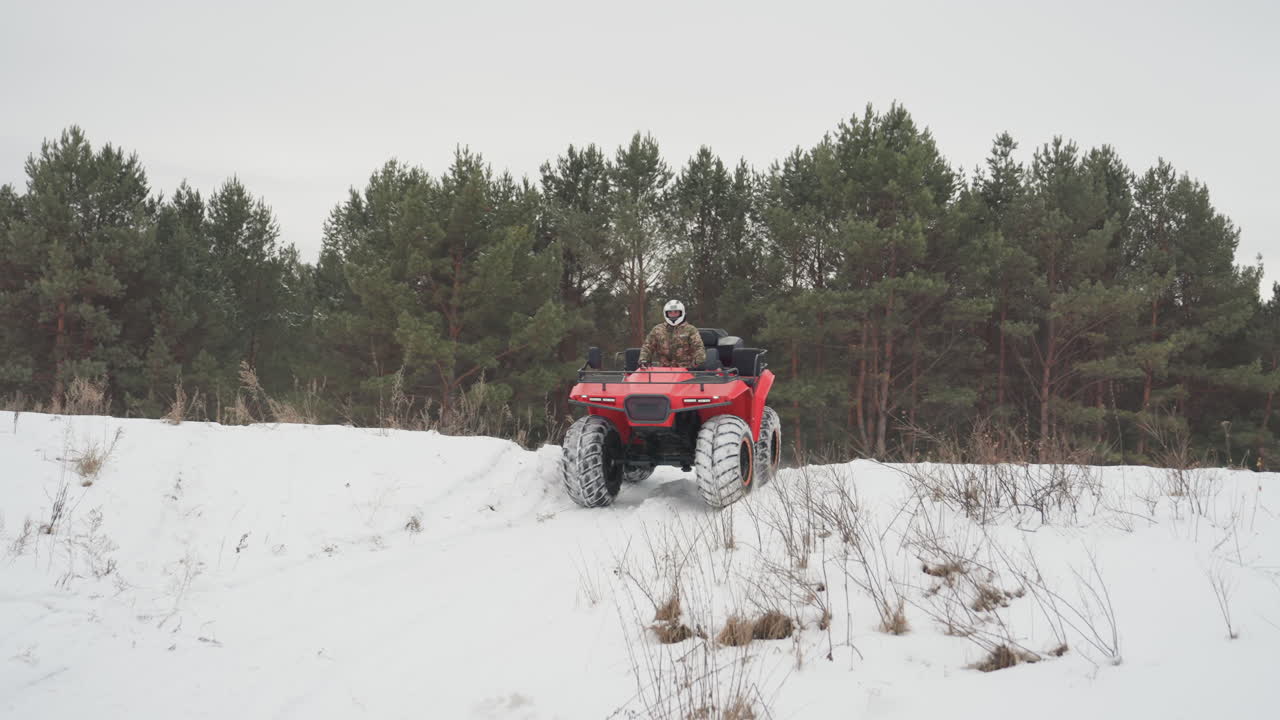 Un ATV rosso si avvicina attraverso un campo innevato. Un pilota bianco, con mimetica e casco, guida costantemente verso la telecamera, mentre le tracce dei pneumatici solcano la neve fresca, con uno sfondo di pini. Una vista frontale ravvicinata che evidenzia.
