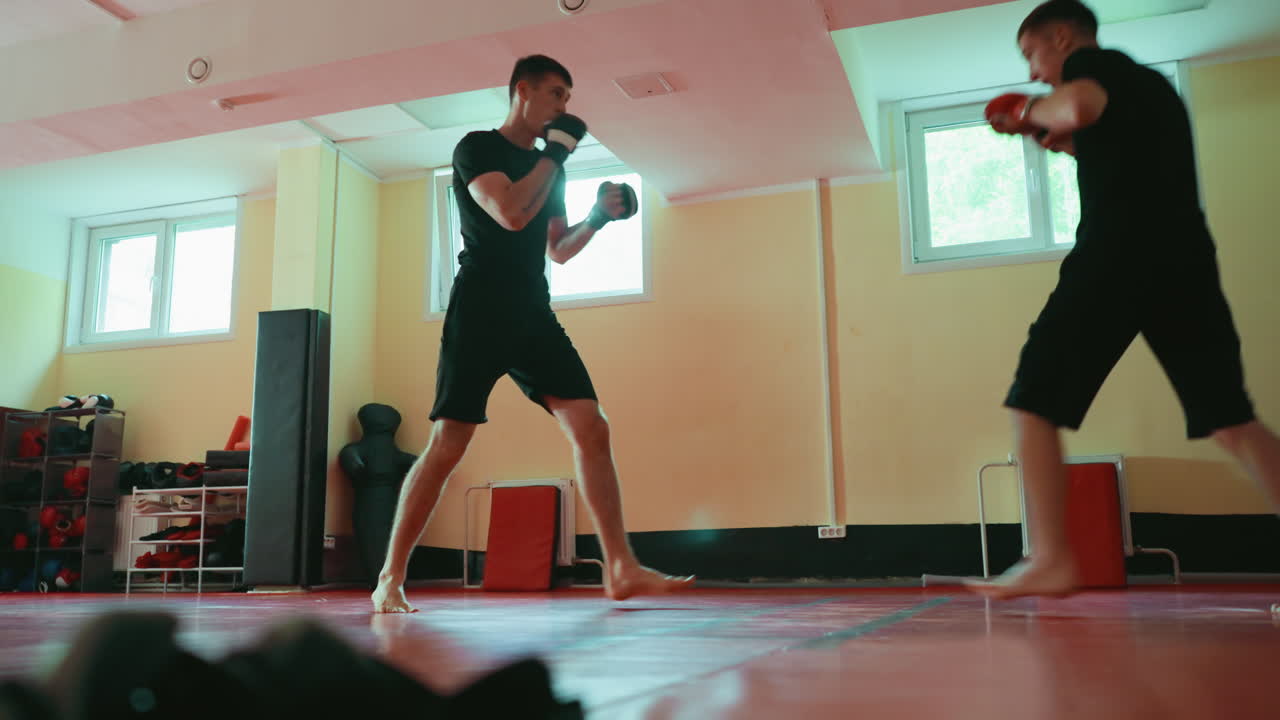 Two fighters sparring in gym on red mat, barefoot, wearing gloves, practicing martial arts techniques, extending punches with focus on combat training