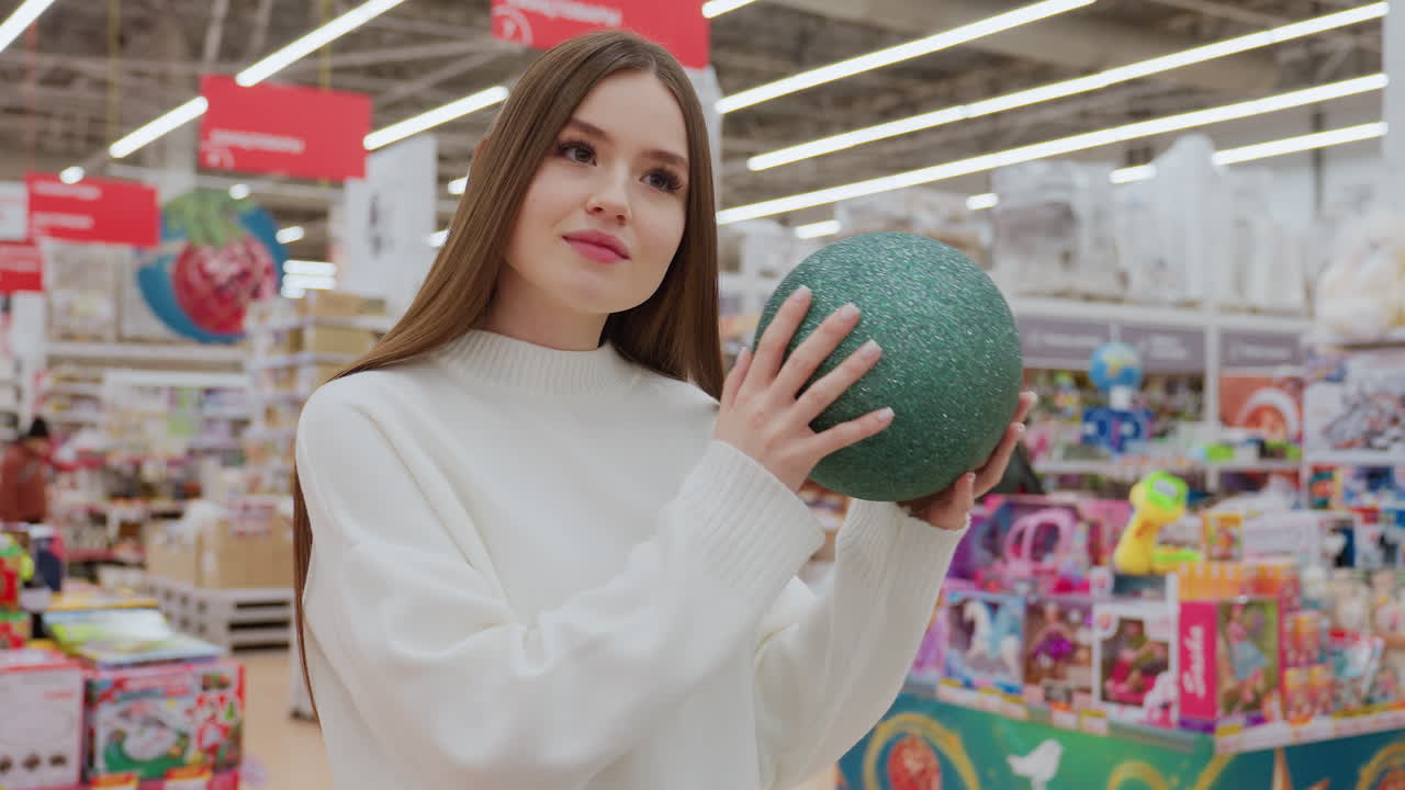 Lady holding green shimmering Christmas ball decor, shaking it near her ear while admiring it in a well-lit decor store with other shoppers in the background