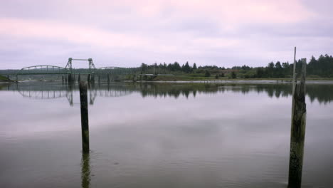 Drone-flying-low-over-Coquille-river-between-two-wooden-poles,-sunrise-pink-colors