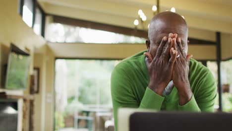 Tired-african-american-senior-man-in-kitchen-working,-using-laptop-and-rubbing-eyes