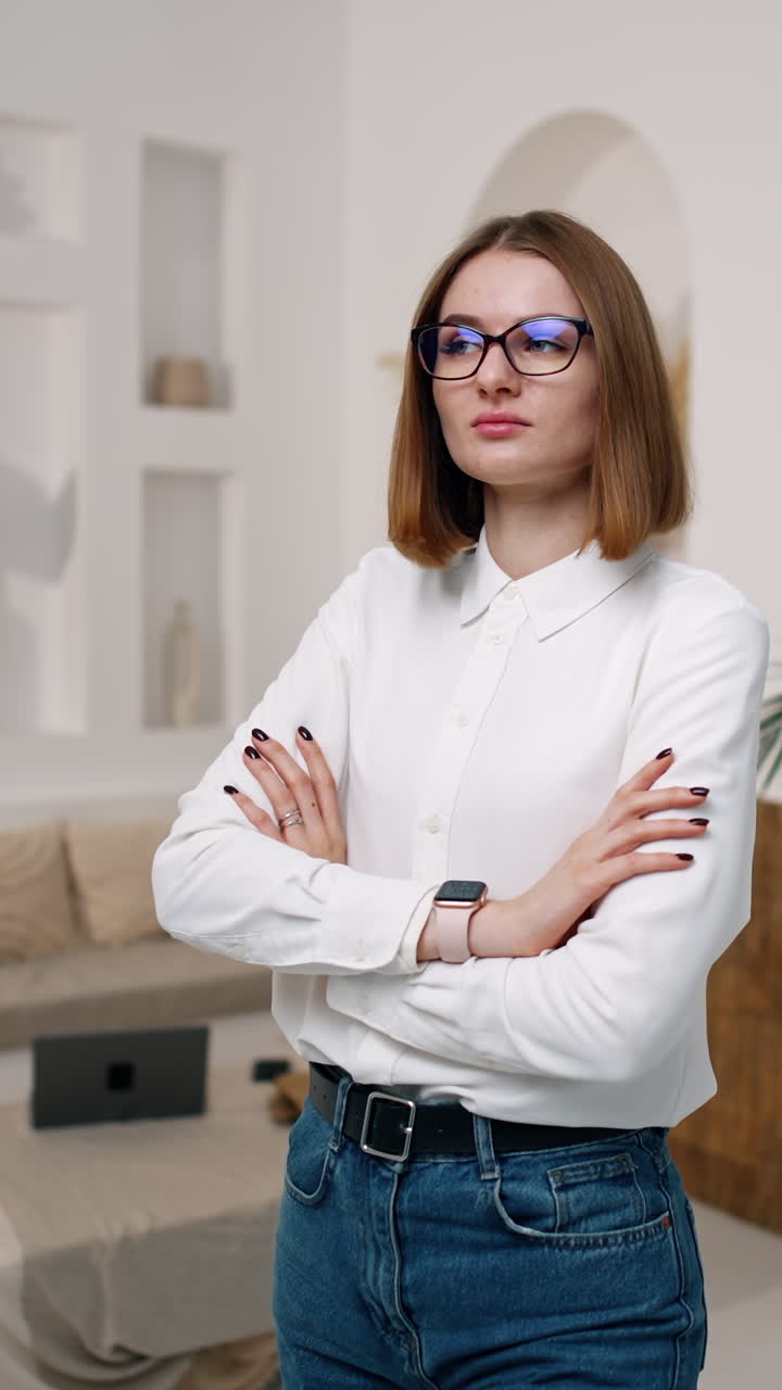 Serious young female touches her hair, fixes her white shirt and crosses her hands on the chest. Portrait of a confident woman looking into camera. Vertical video