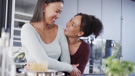 happy biracial lesbian couple preparing food, embracing and laughing in kitchen, in slow motion