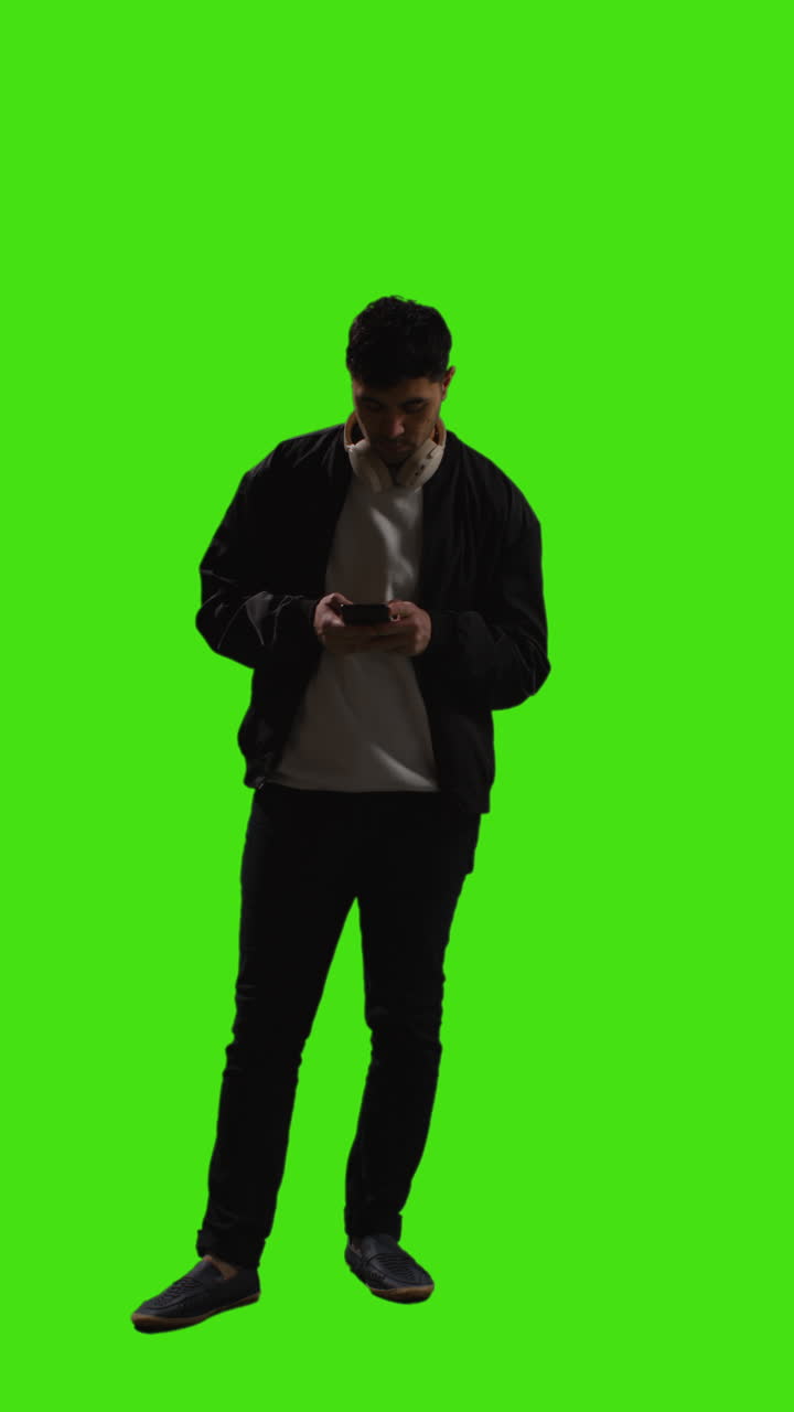 Vertical Video Full Length Shot Of Man Wearing Wireless Headphones Text Messaging On Mobile Phone Against Green Screen