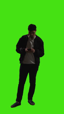 Vertical-Video-Full-Length-Shot-Of-Man-Wearing-Wireless-Headphones-Text-Messaging-On-Mobile-Phone-Against-Green-Screen