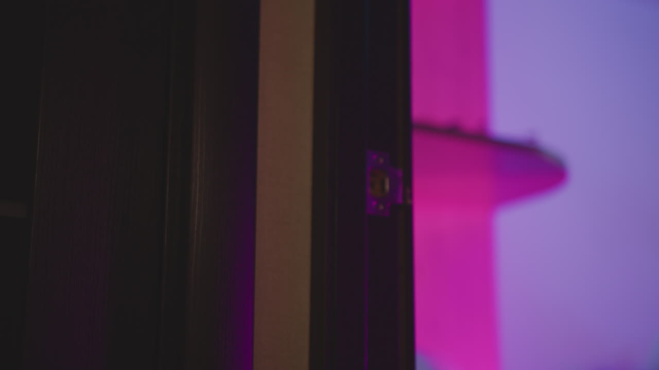 Close up person wearing fluffy sweater opening metal door to neon lit studio bathed in pink purple hue as silhouette hand grip knob and enters vibrant interior under dramatic moody lighting