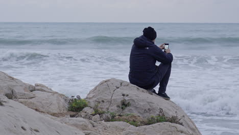 man sitting on a rock taking photos with his phone of stormy sea waves crushing in front of him before departing from the scene