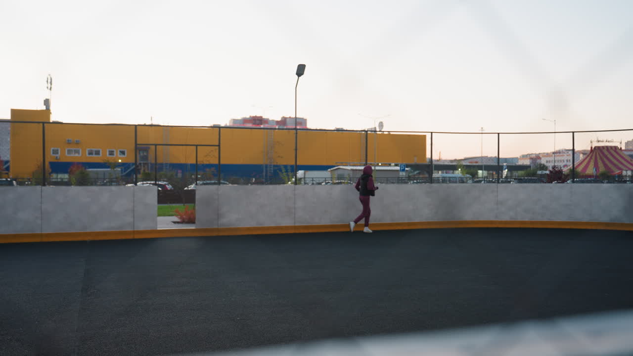 Female jogger runs along curved court under twilight sky, viewed through chain link fence with pastel horizon light and urban backdrop of yellow industrial building and high rise apartments at dusk